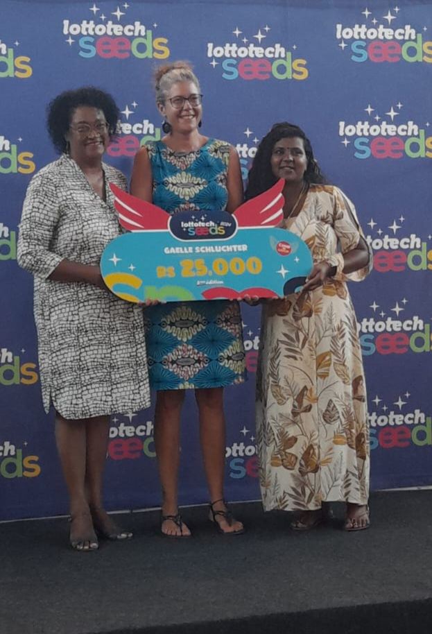 Gaëlle Schluchter reçoit le chèque de 25 000 Rs du prix Lottotech Seeds, entourée de deux membres du jury, devant le mur aux couleurs de Lottotech Seeds